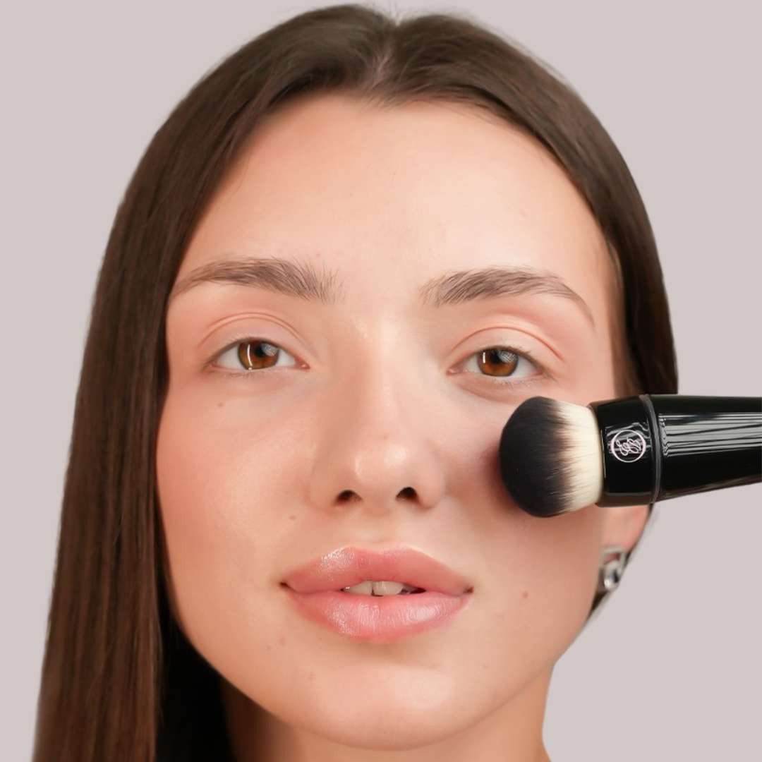 Close-up of a woman applying makeup with a black-handled brush against a plain light gray background.